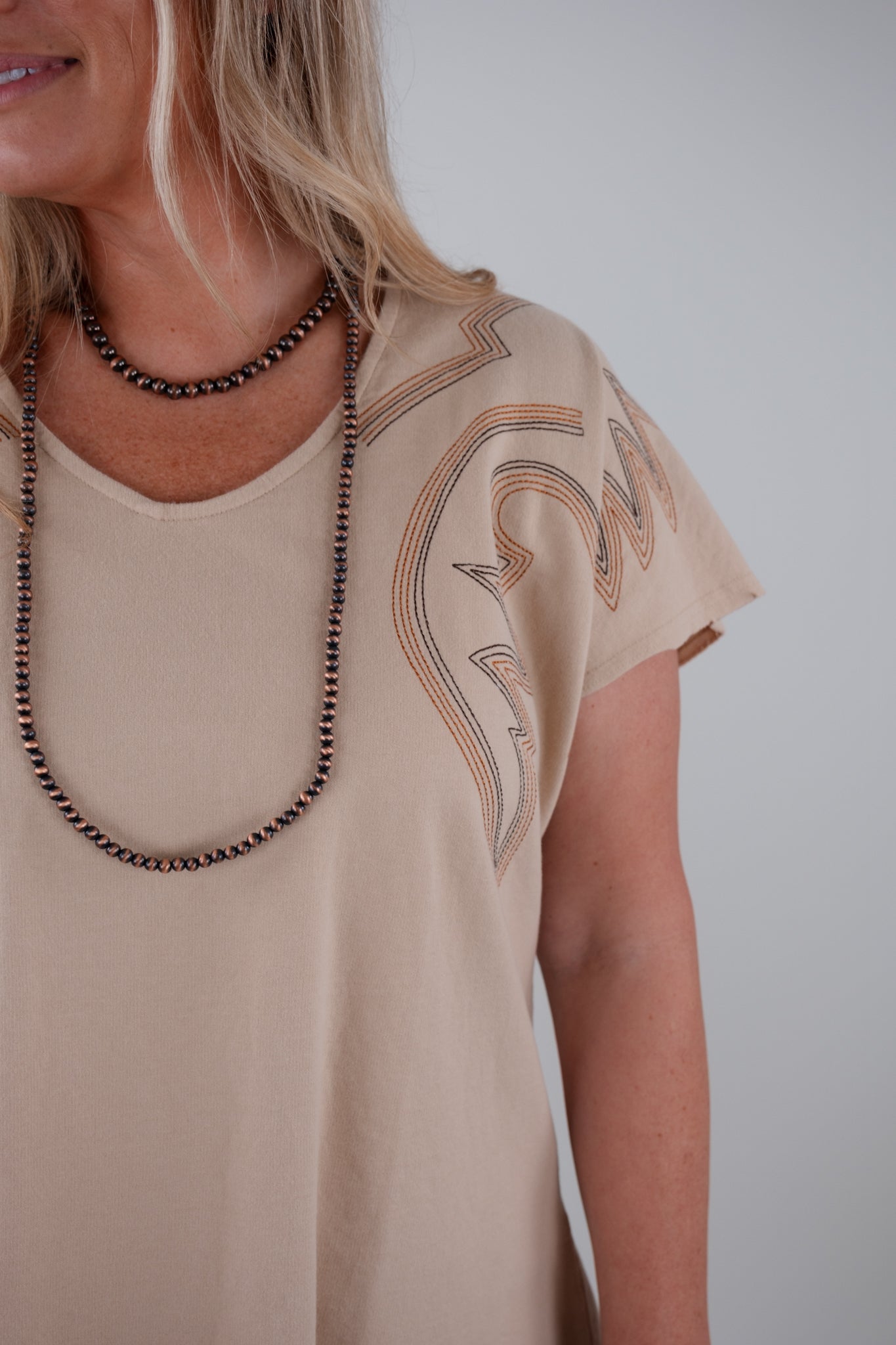 Woman wearing a beige t-shirt with decorative boot stitch patterns and a necklace against a plain background