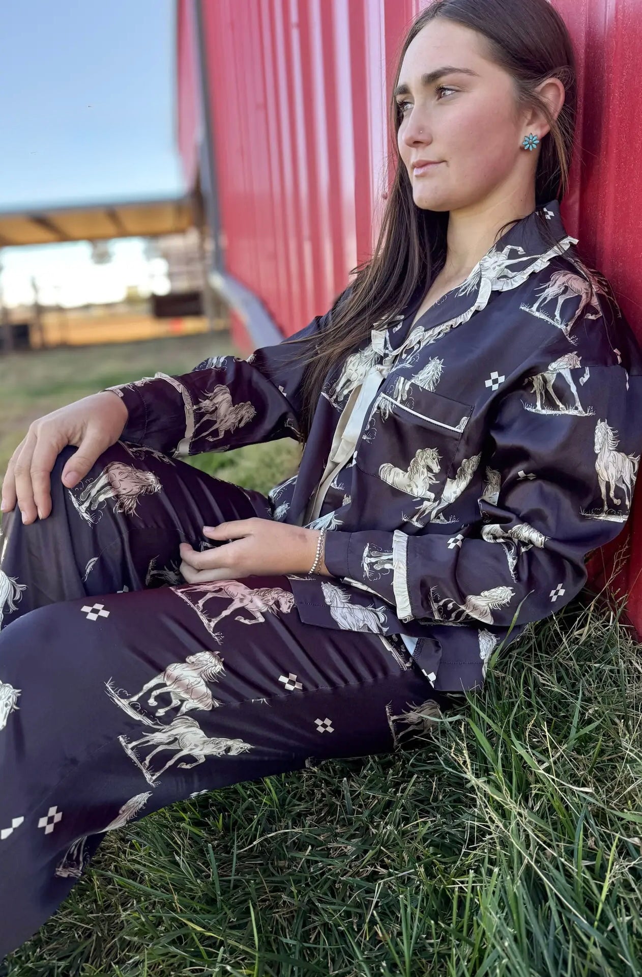 Woman sitting on grass wearing a black pajama set with horse prints against a red barn.