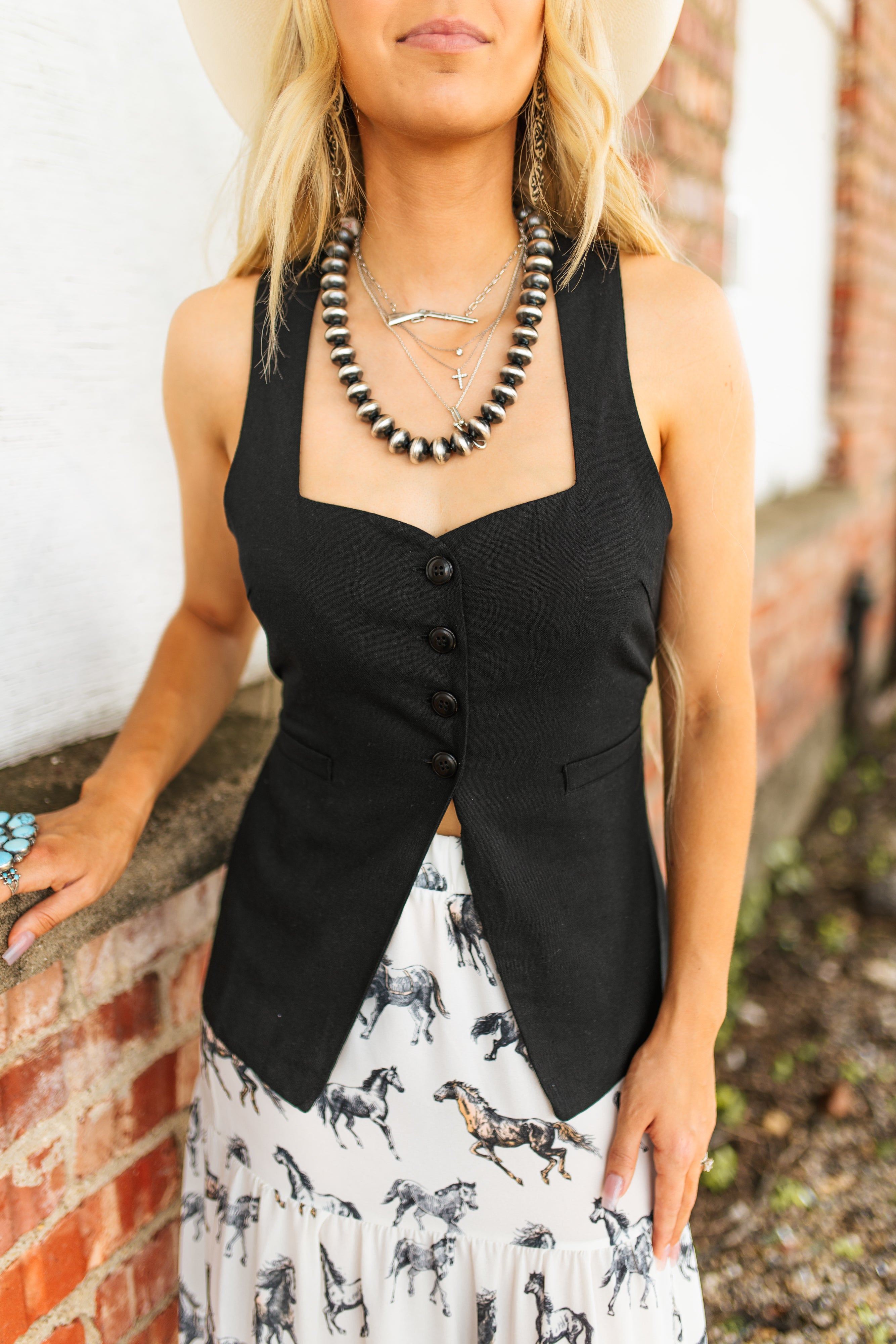 Woman wearing a black vest over a patterned dress with a necklace, standing against a brick wall.