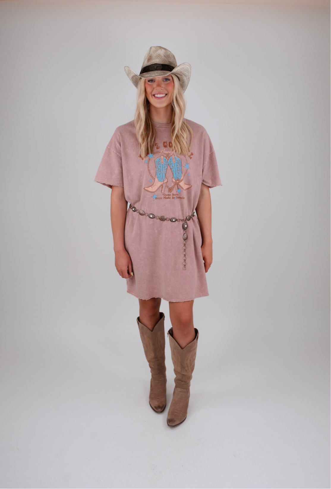 Woman wearing a pink dress with a graphic design, brown boots, and a cowboy hat on a white background