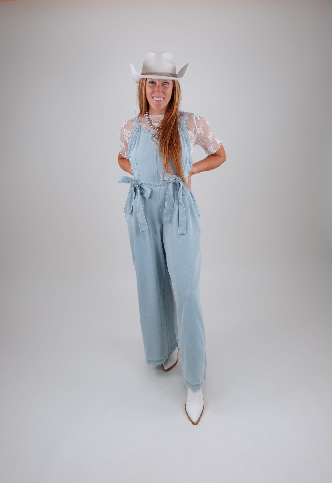 Woman wearing a light blue denim jumpsuit, white cowboy hat, and white boots on a plain background