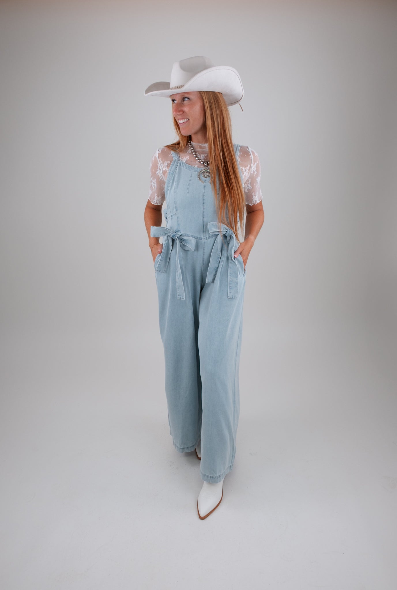 Woman wearing light blue denim overalls and a white cowboy hat on a plain background