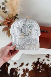 Light camo patterned cap with a horseshoe design held by a hand against a neutral background.
