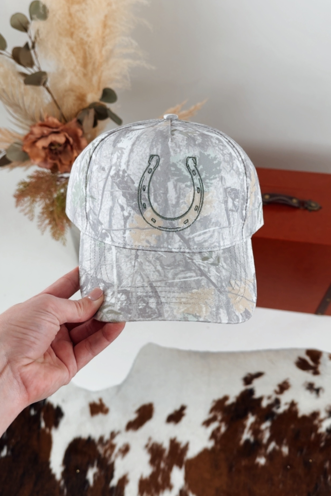 Light camo patterned cap with a horseshoe design held by a hand against a neutral background.