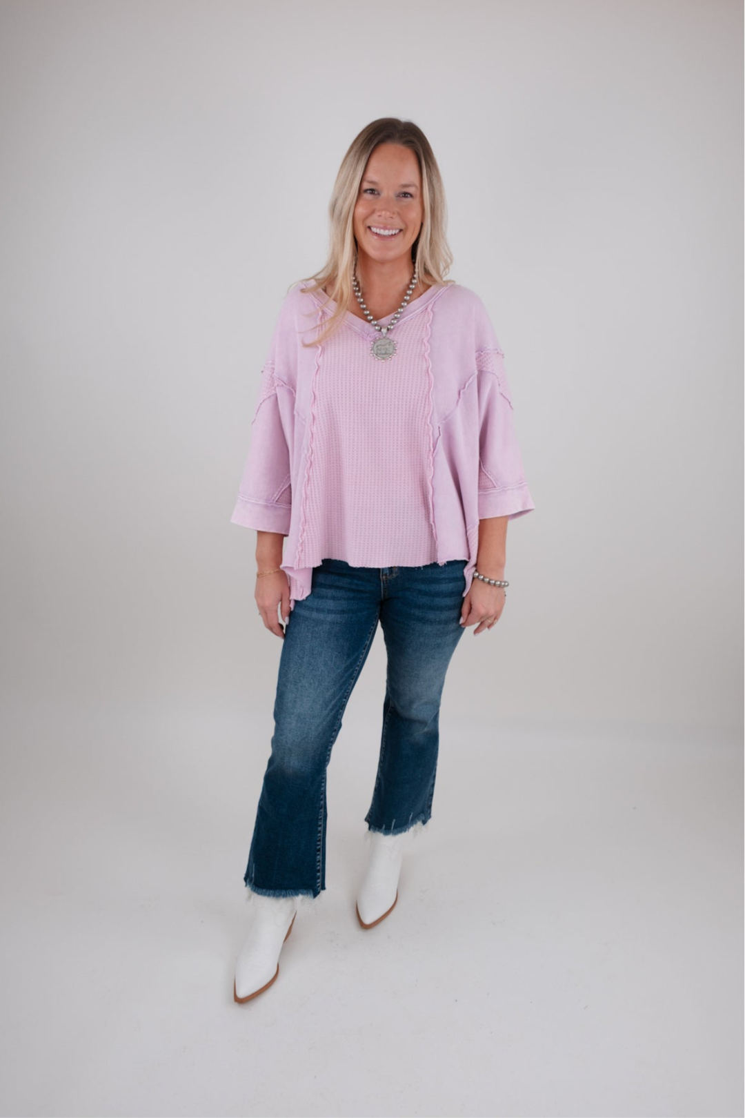 Woman wearing a pink sweater, dark wash blue jeans, and white boots on a plain background