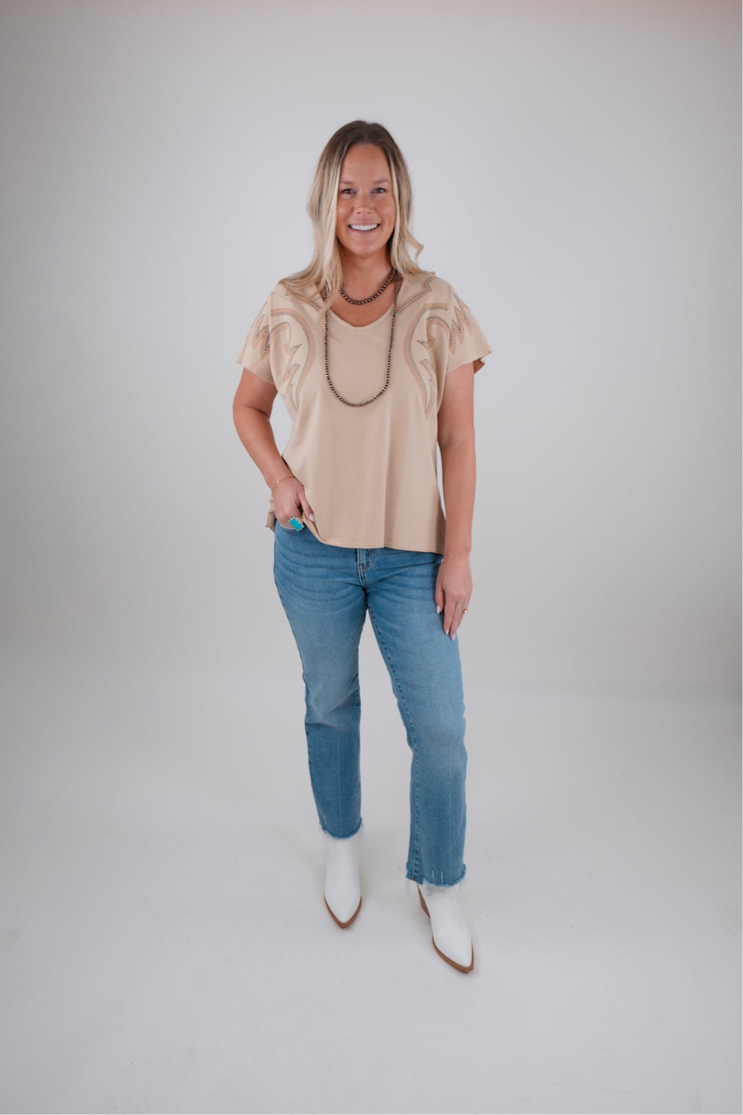 Woman wearing a beige top and blue jeans with white boots on a plain background