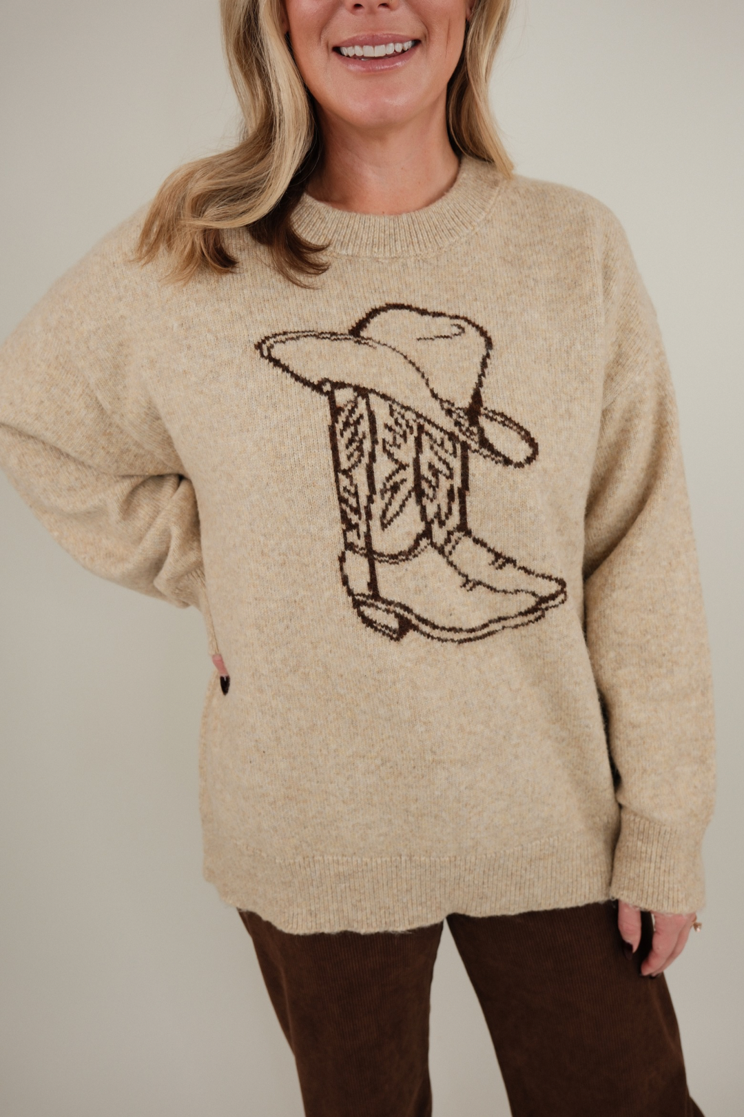 Close-up of Cowgirl Classic Sweater highlighting brown knit design of cowboy hat and boots on soft beige western-inspired fabric.