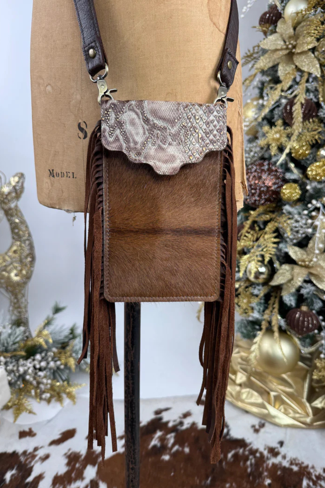 Brown leather crossbody bag with fringe details on a mannequin against a decorated Christmas tree background.