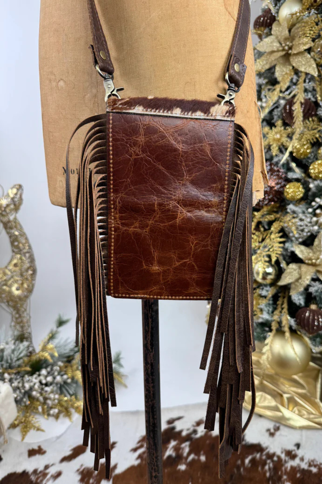 Brown leather crossbody bag with fringes on a mannequin against a Christmas-themed background.