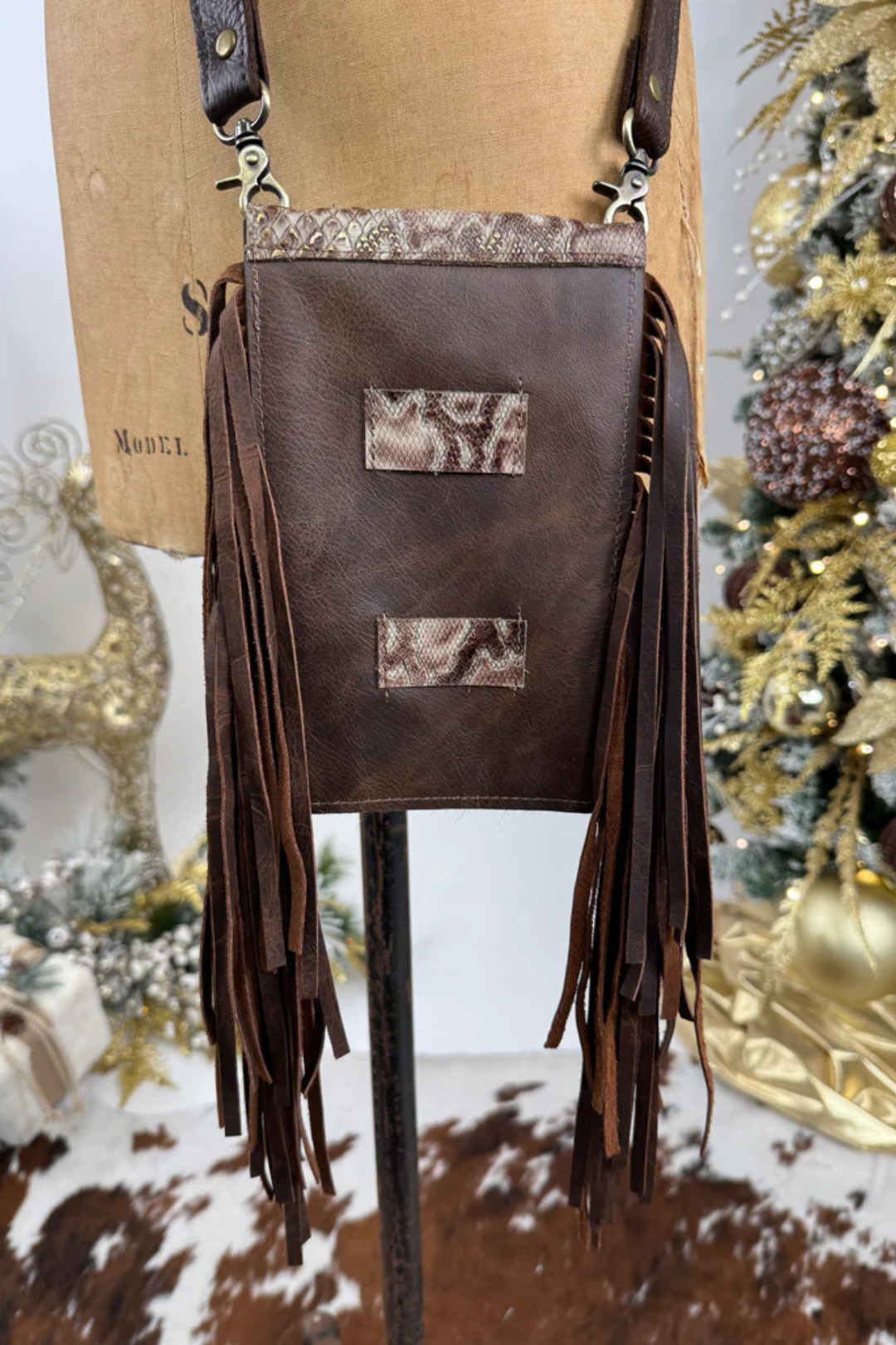 Brown leather crossbody bag with fringe details on a stand against a decorated background.