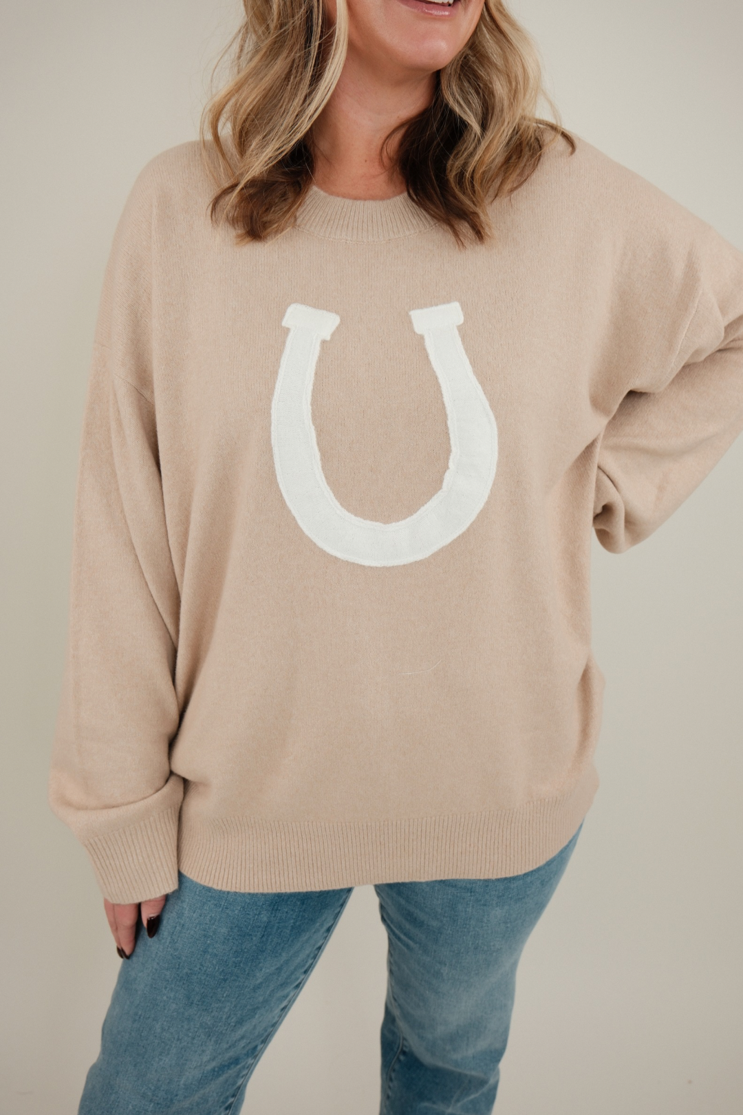 Model wearing beige Cowgirl’s Luck Sweater styled with denim, ultra-soft neutral knit with horseshoe design for cozy western chic.