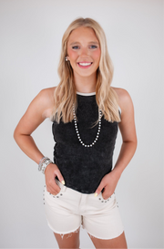 Woman wearing a black sleeveless top and white shorts with a pearl necklace on a plain background