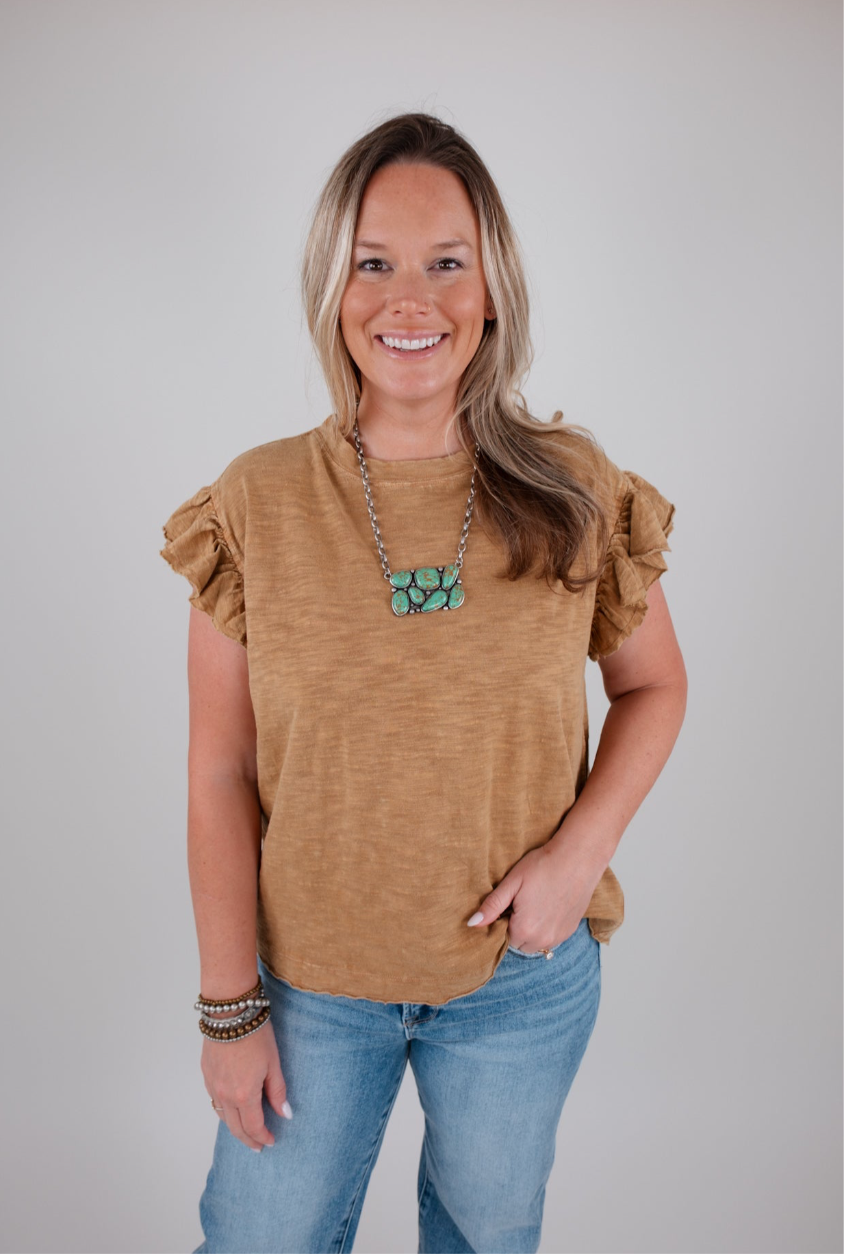 Woman wearing a camel colored ruffled top and blue jeans against a plain background