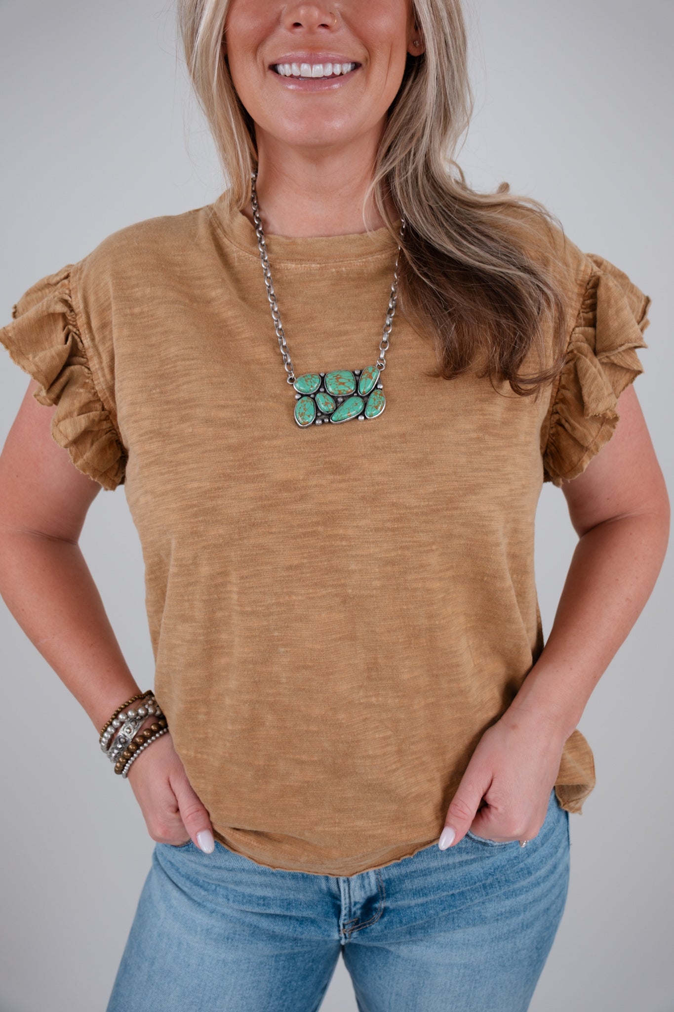 Woman wearing a camel colored ruffled shirt and blue jeans with a gray background