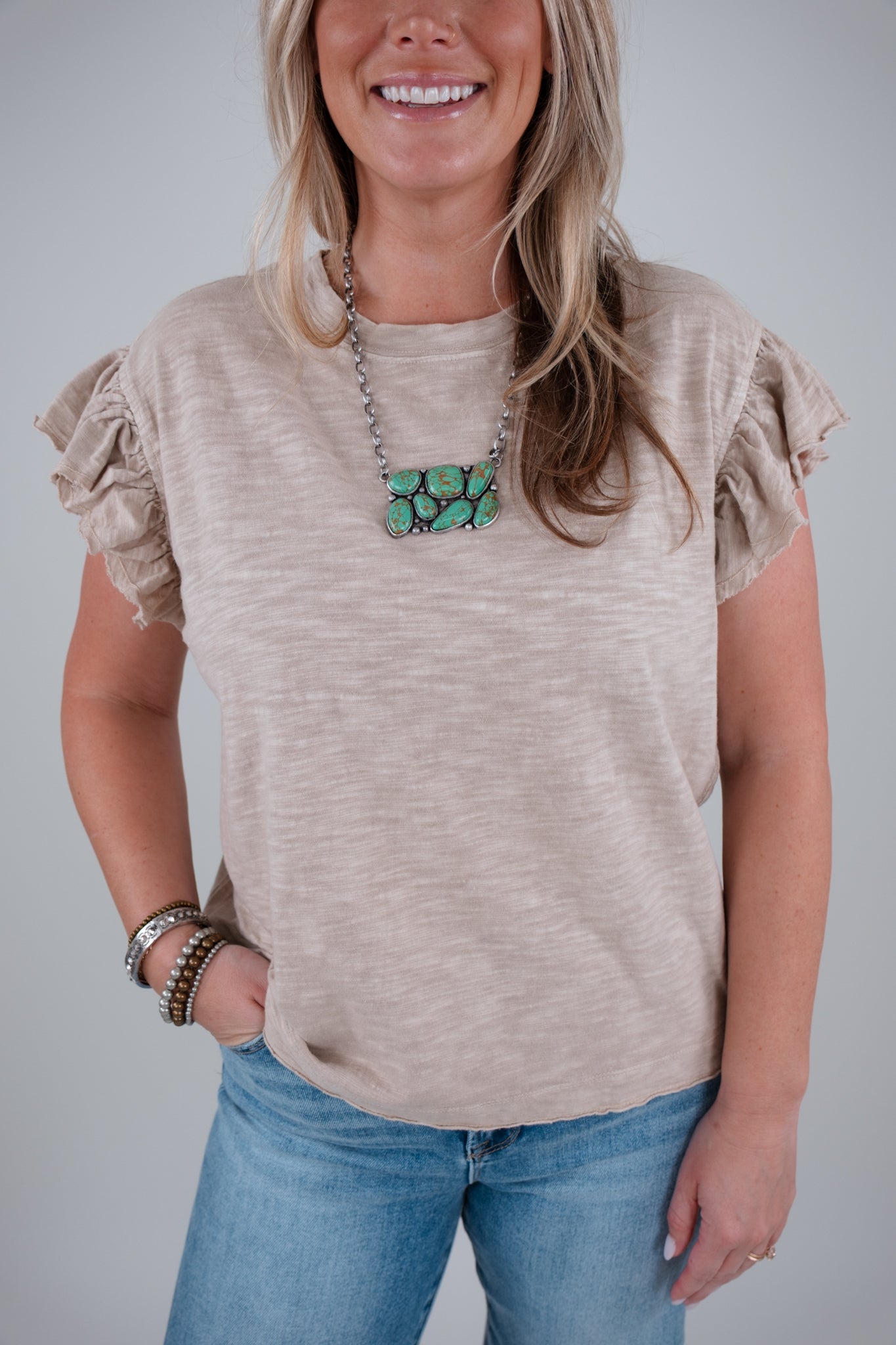 Woman wearing a beige top with ruffled sleeves and blue jeans, smiling against a plain background.