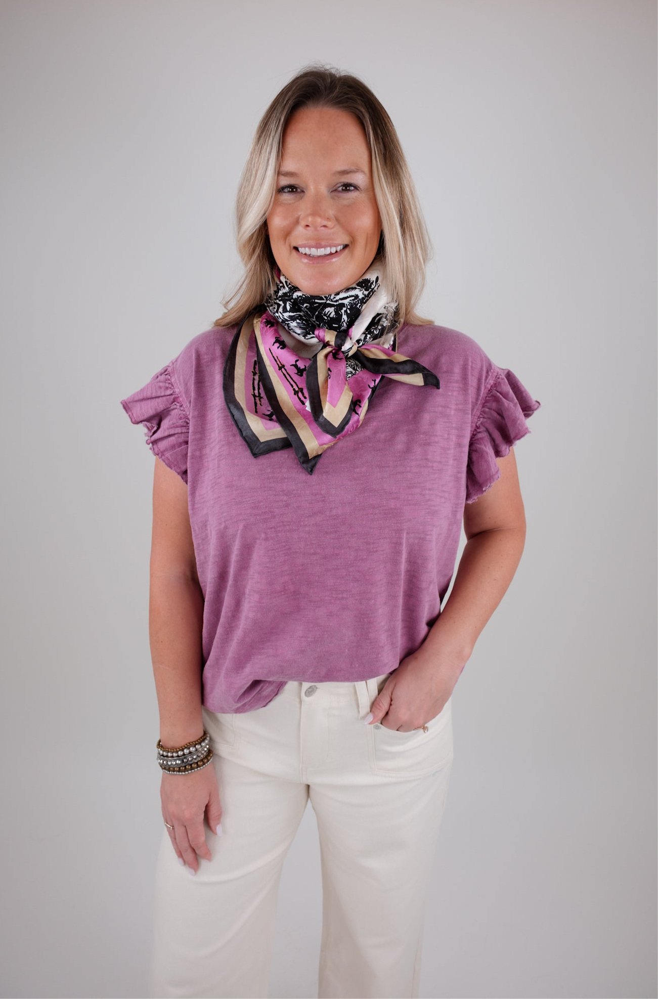 Woman wearing a purple top and white pants with a patterned scarf around her neck on a plain background