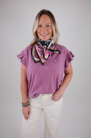 Woman wearing a purple top and white pants with a patterned scarf around her neck on a plain background