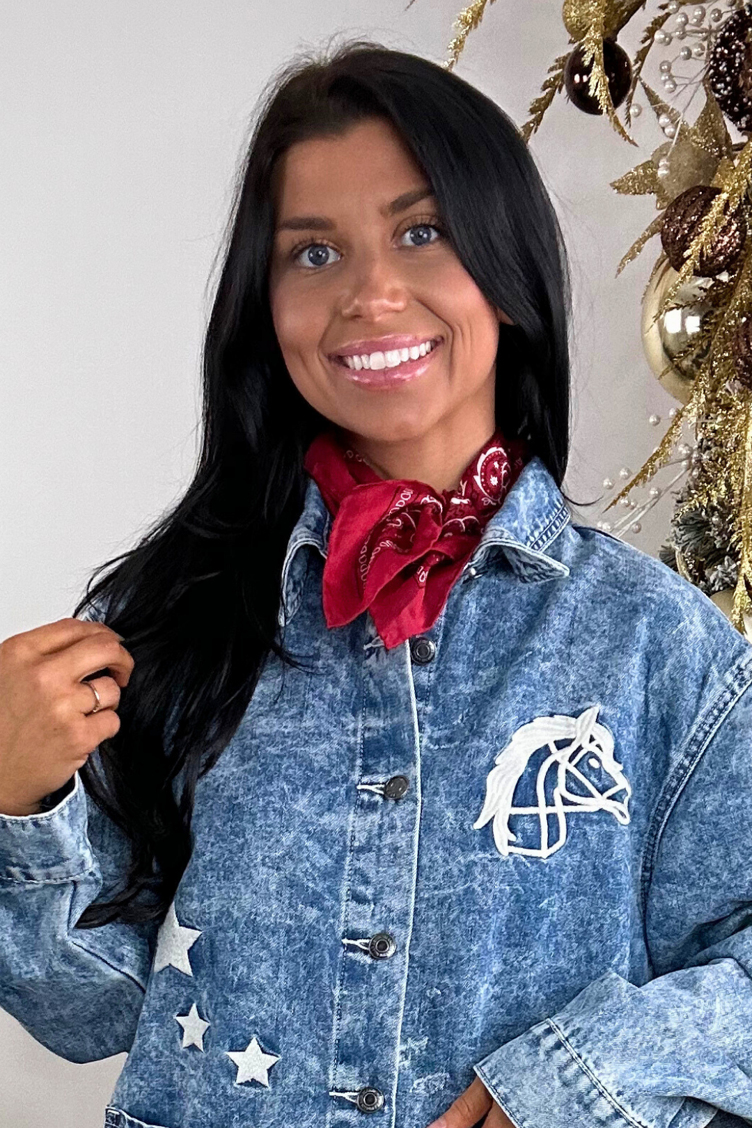 Woman wearing a denim jacket with a horse emblem and a red bandana, standing against a neutral background.