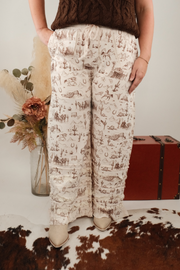 Person wearing western patterned pants standing next to a vintage suitcase and floral arrangement.
