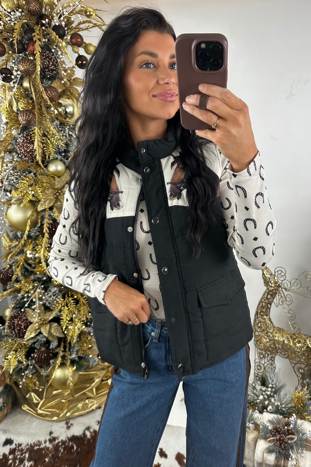 Woman wearing a horse vest taking a mirror selfie in front of a decorated Christmas tree, wearing a black vest over a patterned shirt.