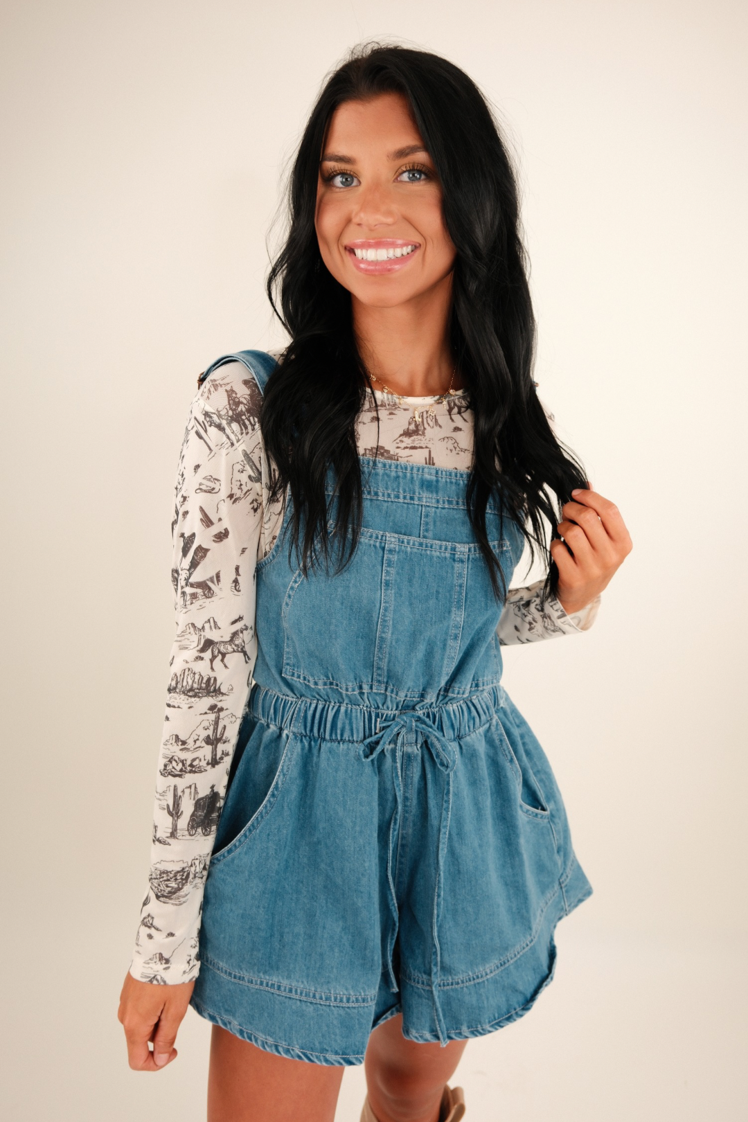 A model in a western‑print mesh long‑sleeve top, featuring a round neckline and tonal cactus, cowboy hat, boot and horse head design on sheer black fabric.