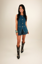 A model wearing a dark wash denim vest with gold buttons down the front, chest flap pockets, and Western yoke detailing.