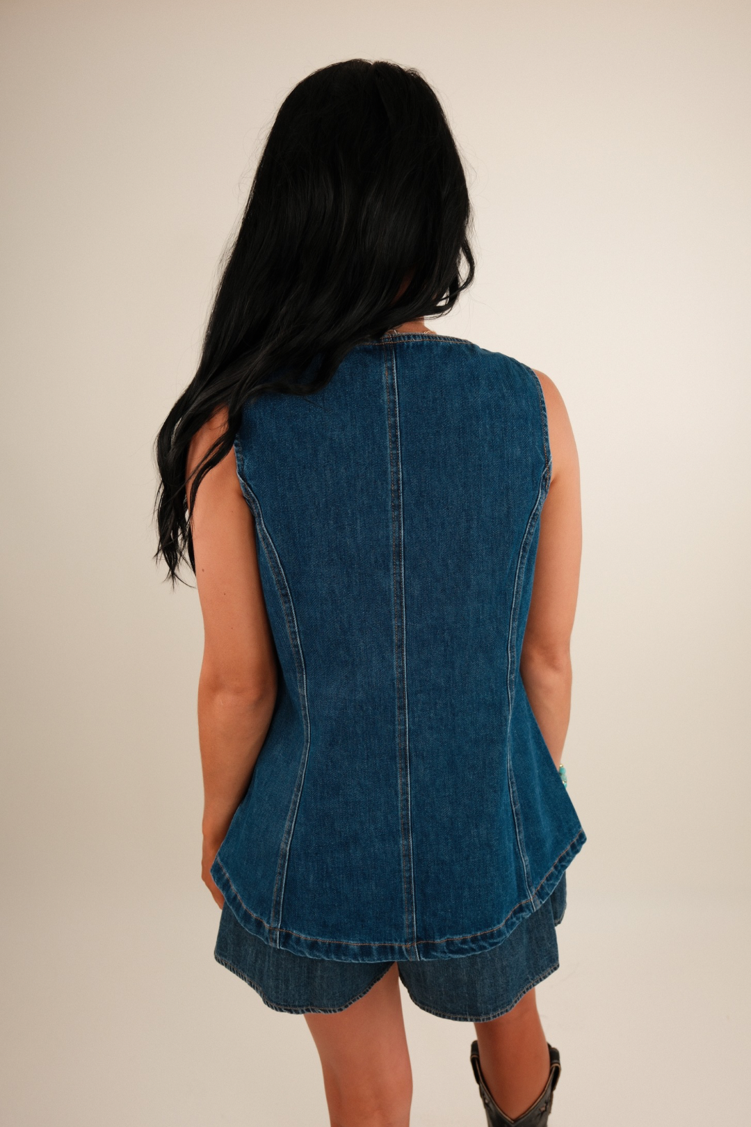Back of the dark wash denim vest showing a Western‑style yoke and raw hem edge with a relaxed fit.