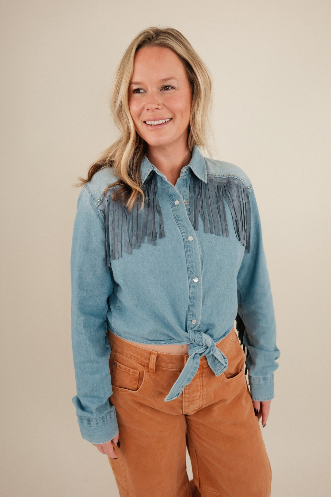 Front view of High Noon Fringe Denim Top, cropped light wash button-down with long fringe across chest and sleeves, snap closure and tie waist.