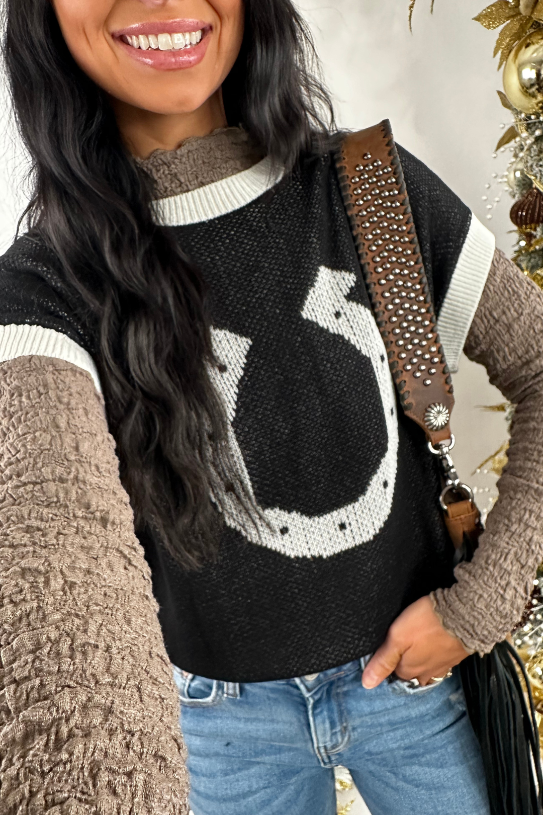 Woman wearing a black sweater with a horseshoe design, standing next to a decorated Christmas tree.