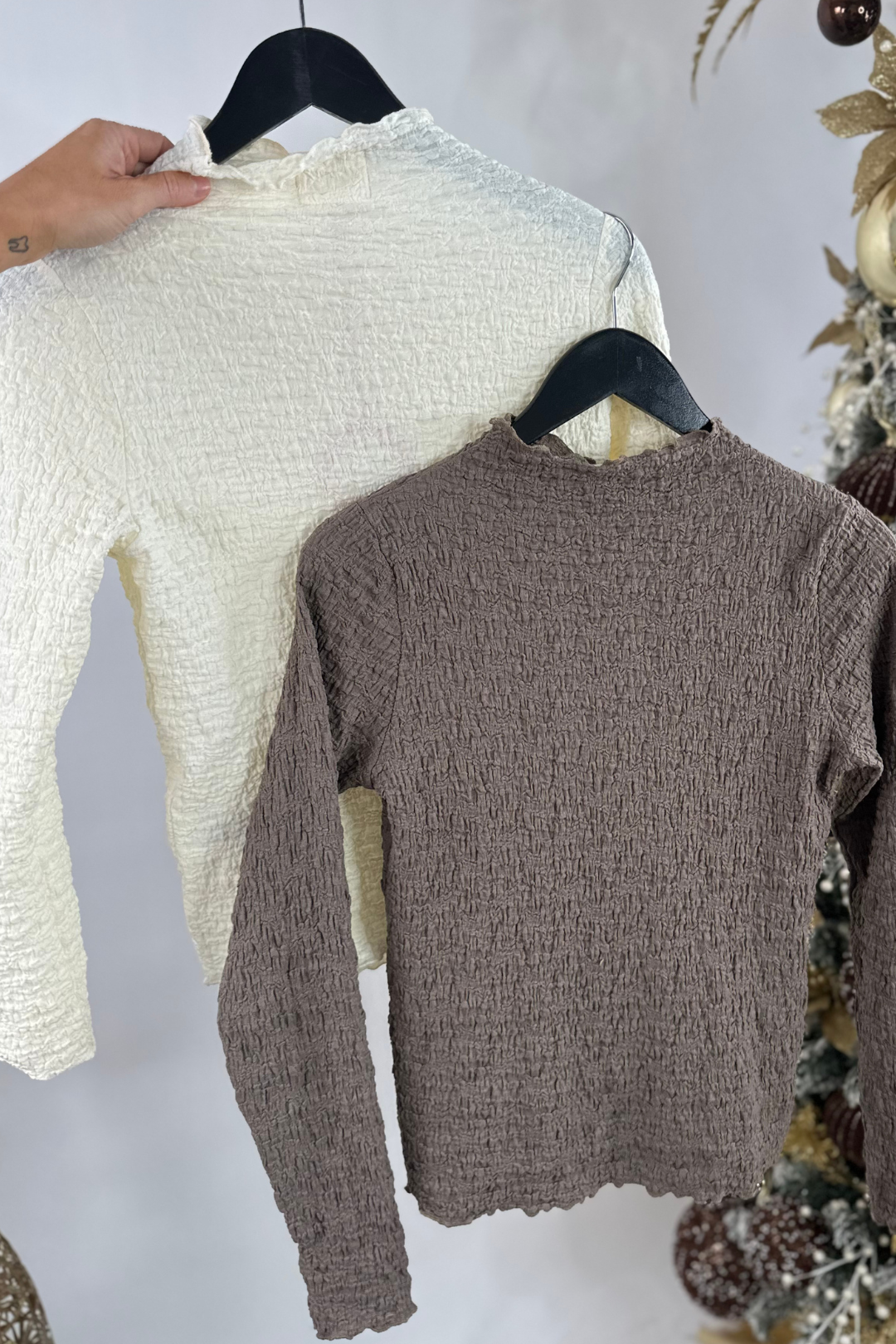 Two long sleeve blouses, one ivory and one taupe, on hangers against a white background.