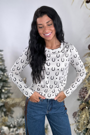 Woman wearing a white long-sleeve top with black horseshoe pattern and blue jeans against a decorative background.