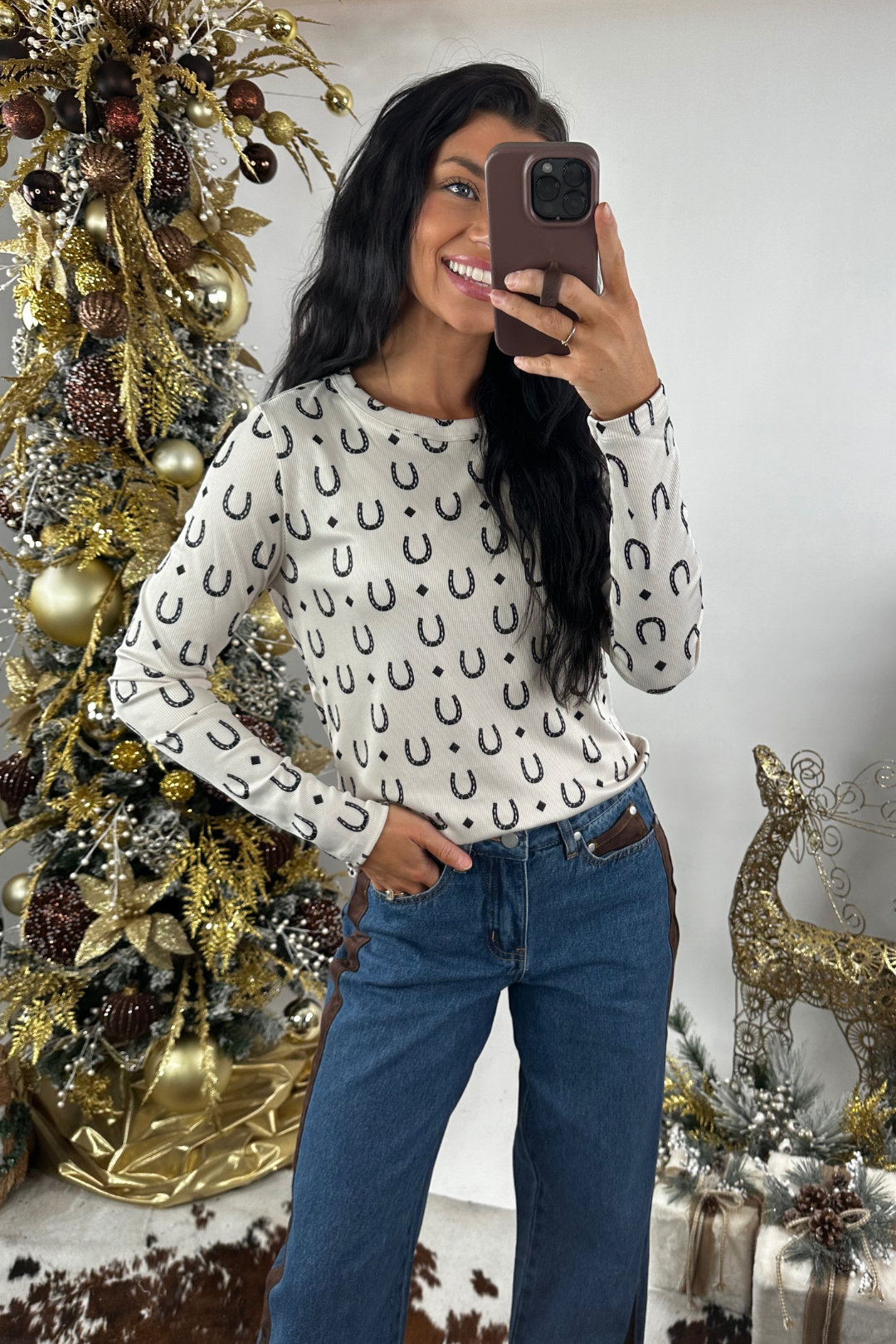 Person wearing a white long sleeve top with black horseshoe pattern taking a mirror selfie in front of a decorated Christmas tree