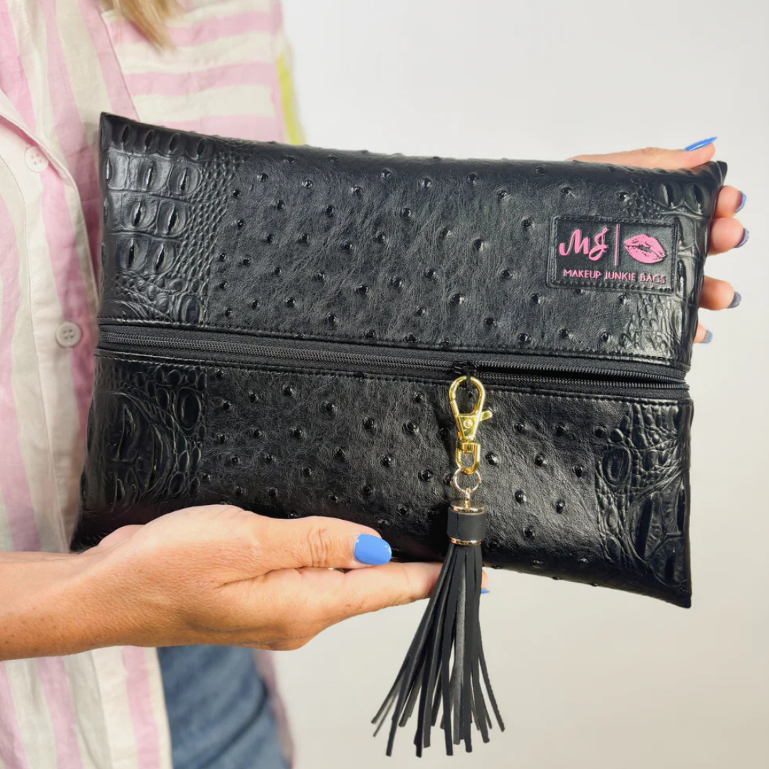 Person holding sleek Obsidian Makeup Junkie bag in black vegan leather, showing gold tassel zipper detail and compact western glam style.