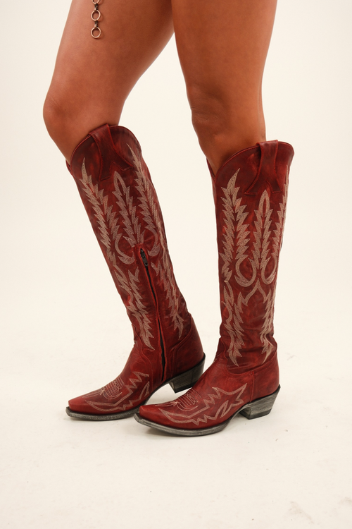 Old Gringo Red Mayra Boots - A pair of tall distressed-red leather cowboy boots standing on a rock outdoors, showcasing ivory western-style embroidery on the shaft and vamp, pointed snip toes, and a riding-style heel.