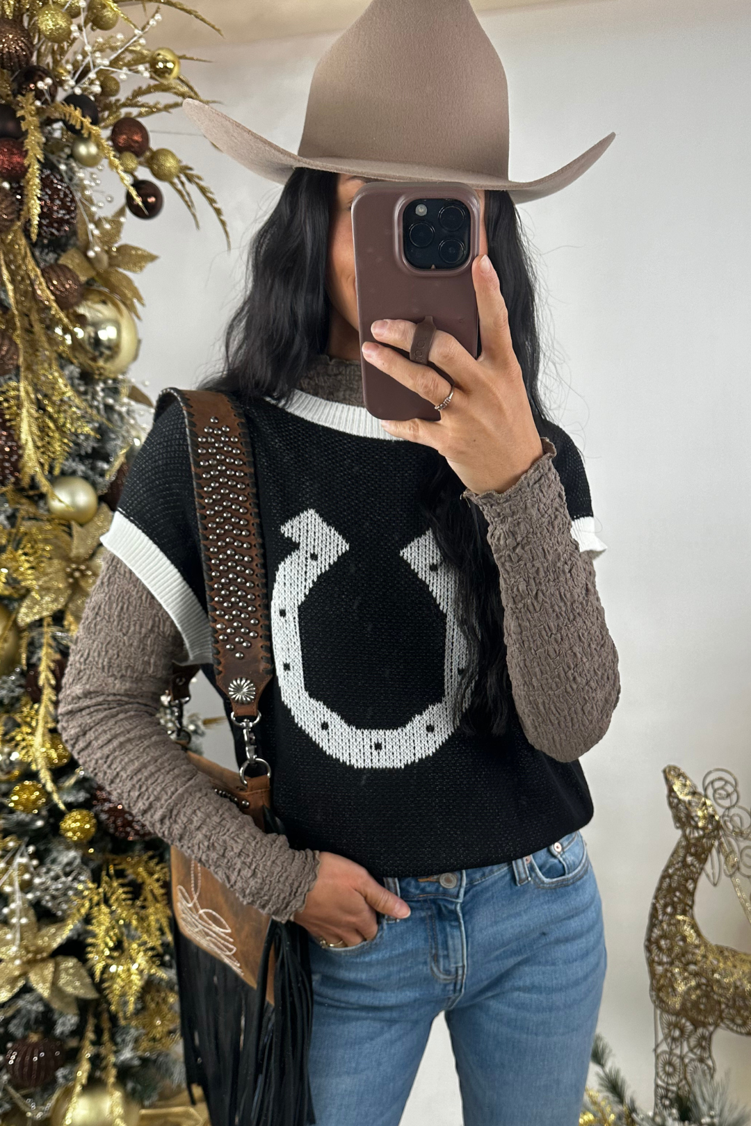 Person wearing a black sweater with a white horseshoe design, holding a phone, standing in front of a decorated Christmas tree.