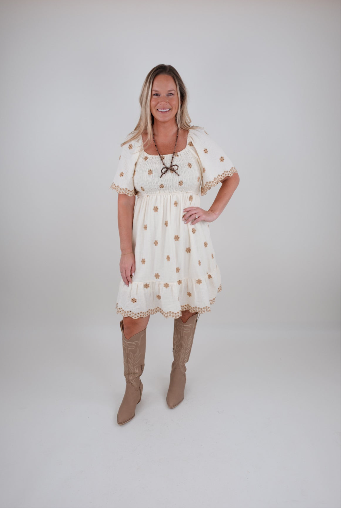 Woman wearing a white dress with gold eyelet flowers and brown knee-high boots on a plain background
