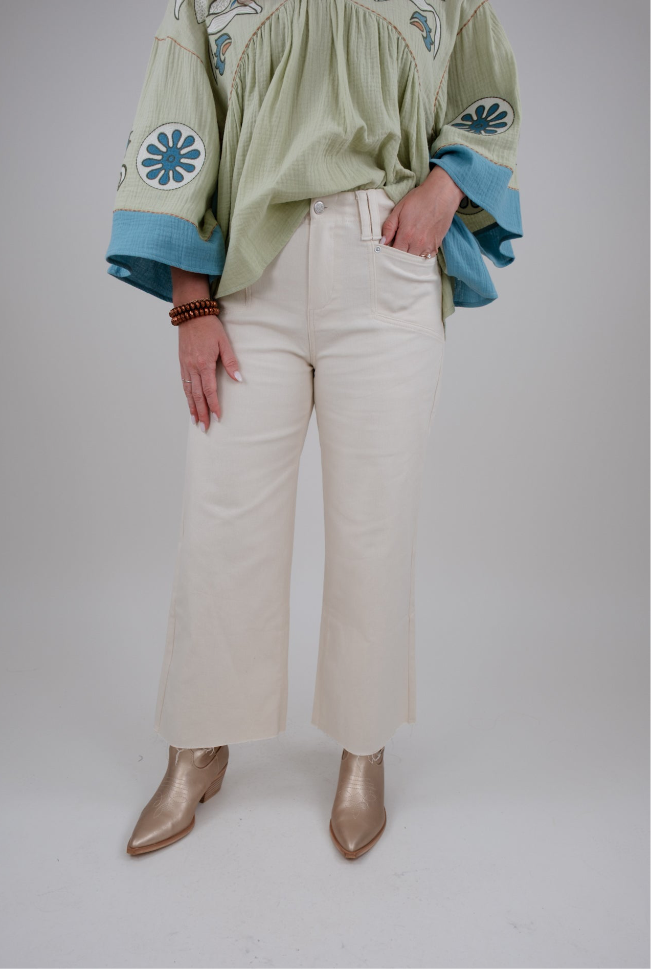 Person wearing a green embroidered blouse with blue details, beige pants, and gold boots on a white background