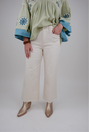 Person wearing a green embroidered blouse with blue details, beige pants, and gold boots on a white background