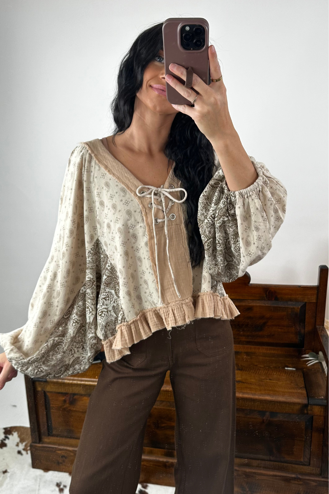 Woman taking a mirror selfie wearing a patterned blouse and brown pants.