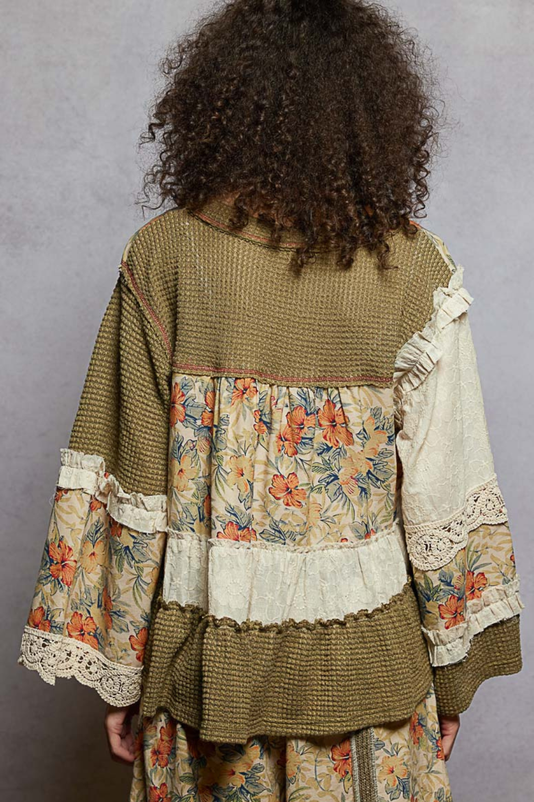Back side of a woman wearing a patchwork blouse with floral and lace details on a gray background