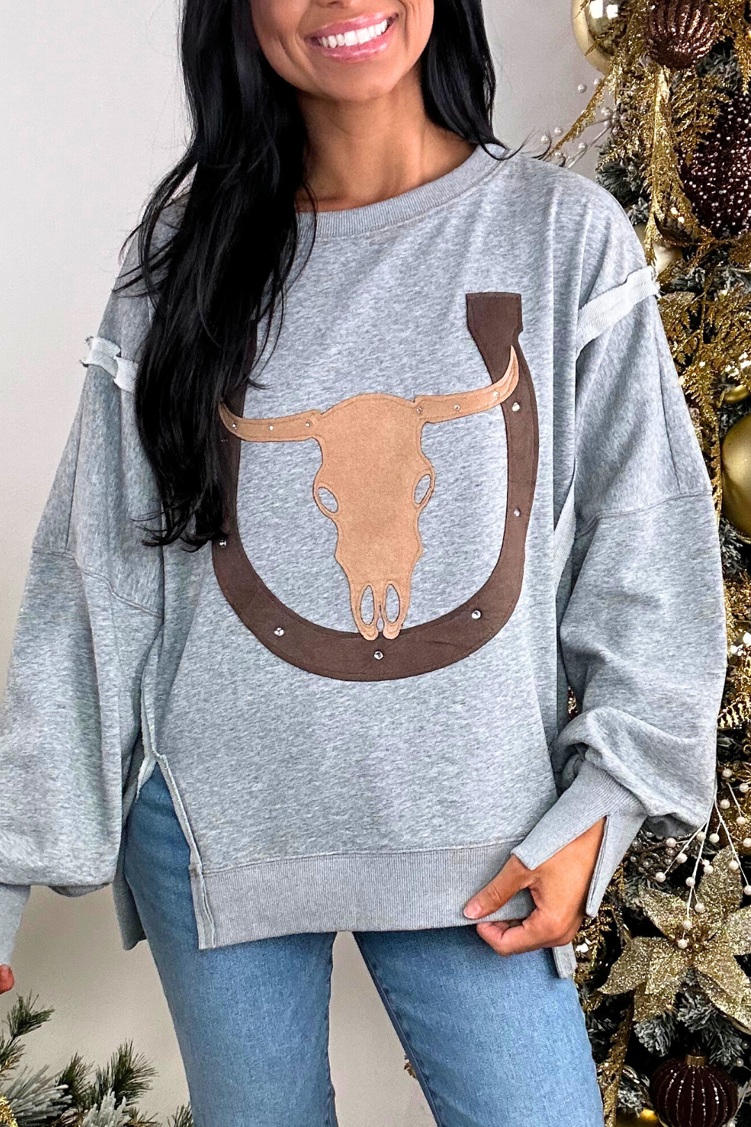 Person wearing a gray sweatshirt with a bull skull design, standing next to a decorated Christmas tree.