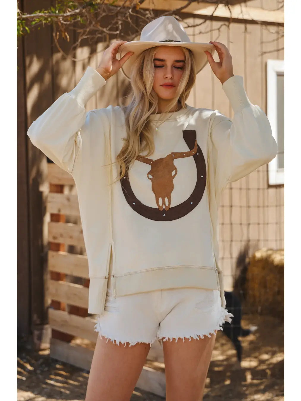 Woman wearing a white sweatshirt with a horse and horseshoe design outdoors.