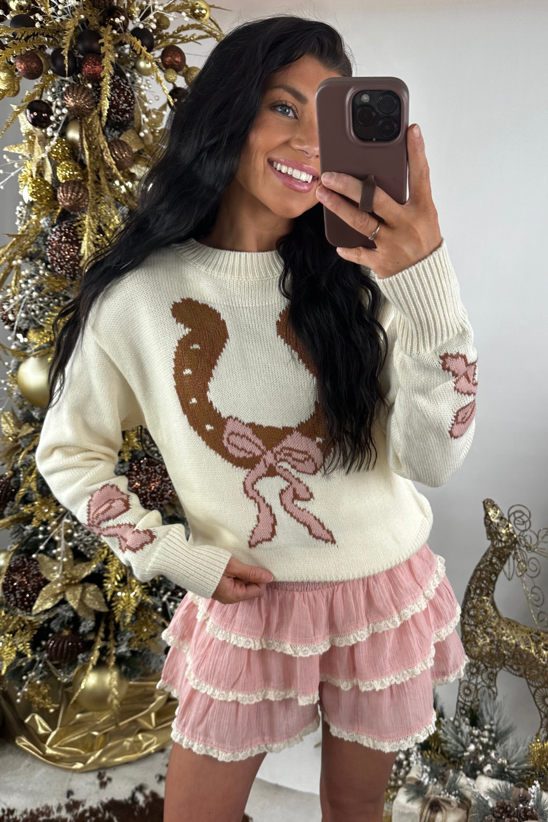 Woman wearing a cream sweater with a horseshoe design and pink skort, taking a selfie in front of a decorated Christmas tree.