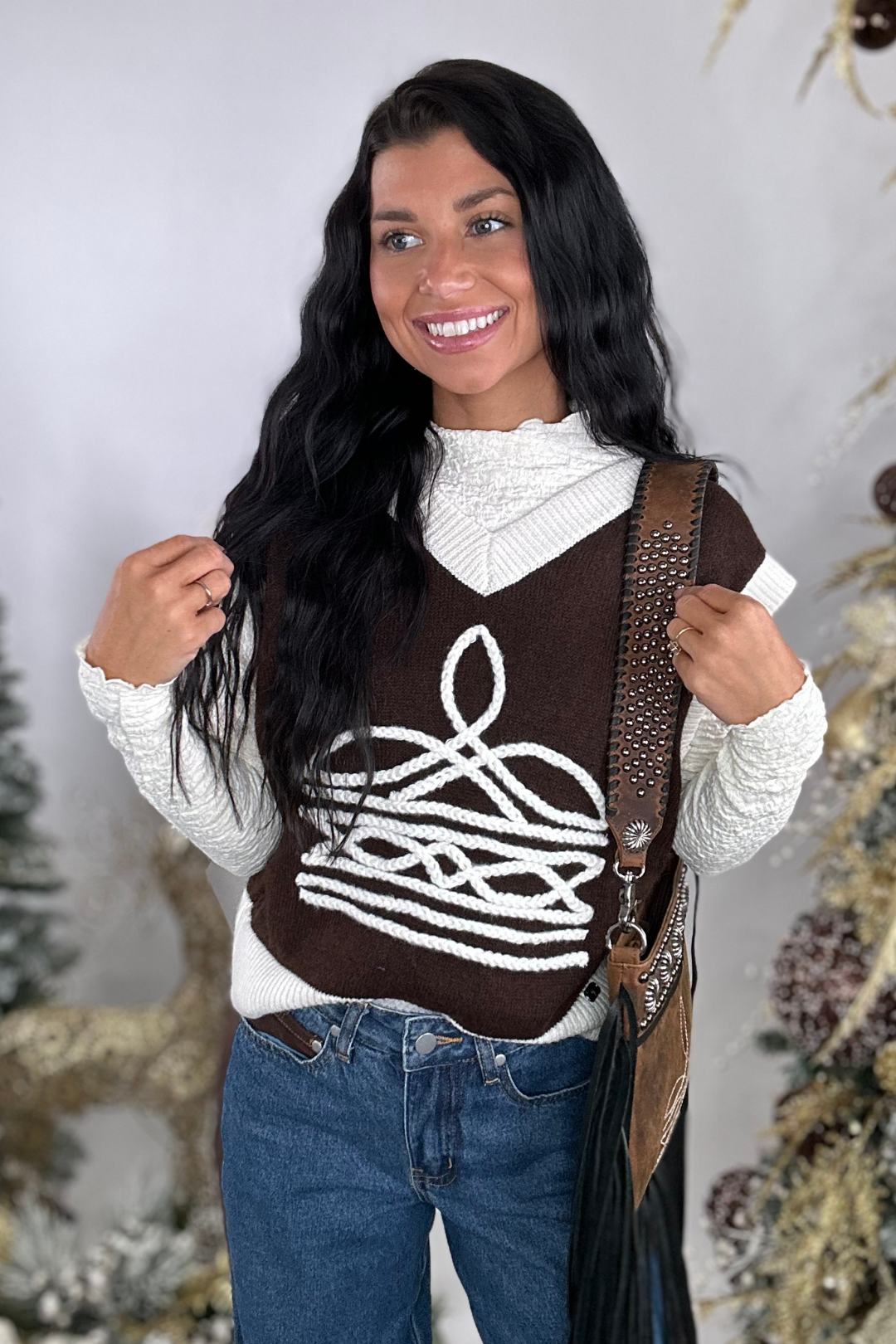 Woman wearing a brown sweater vest with a white pattern, white turtleneck, blue jeans, and carrying a brown bag.