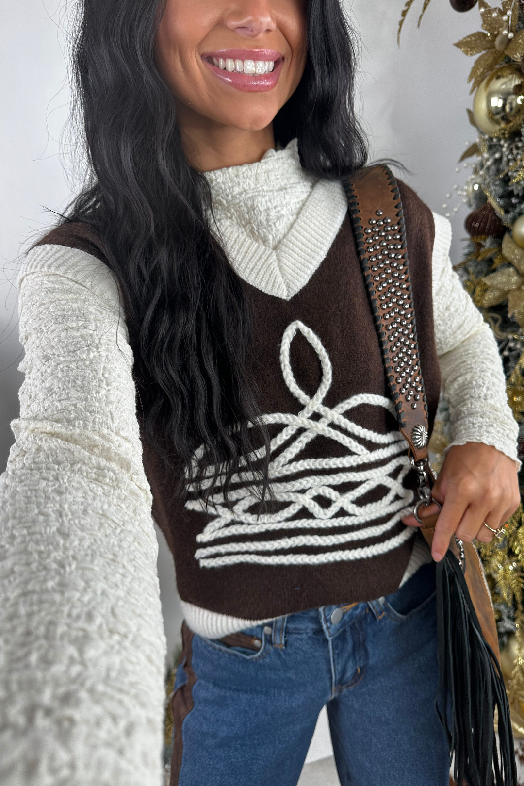 Woman wearing a brown patterned vest over a white sweater, standing in front of a decorated Christmas tree.