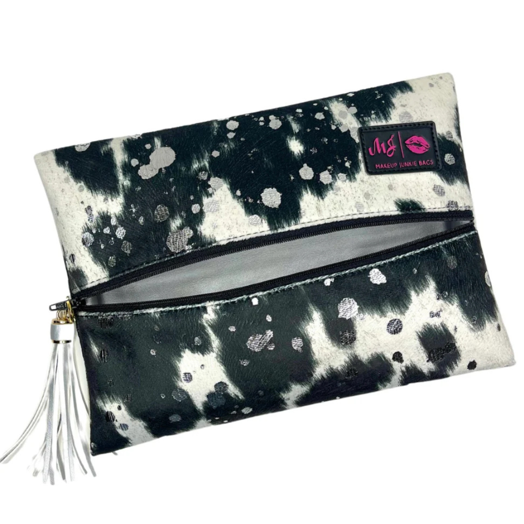 Front view of Rodeo Queen Makeup Junkie Bag with black‑and‑white vegan cowhide print, acid‑wash sparkle, black zipper and tassel.