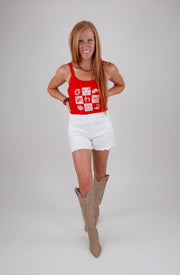 Woman wearing a red tank top with white patterns and white shorts, standing against a plain background.