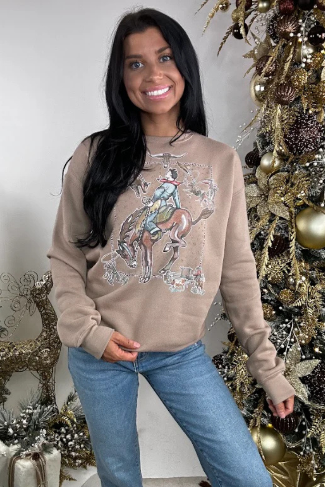 Woman wearing a beige sweatshirt with a western graphic design in front of a decorated Christmas tree.