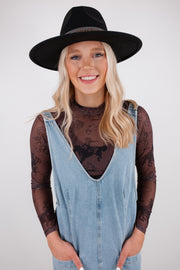 Person wearing a black hat, denim overalls, and sheer sleeves with a white background