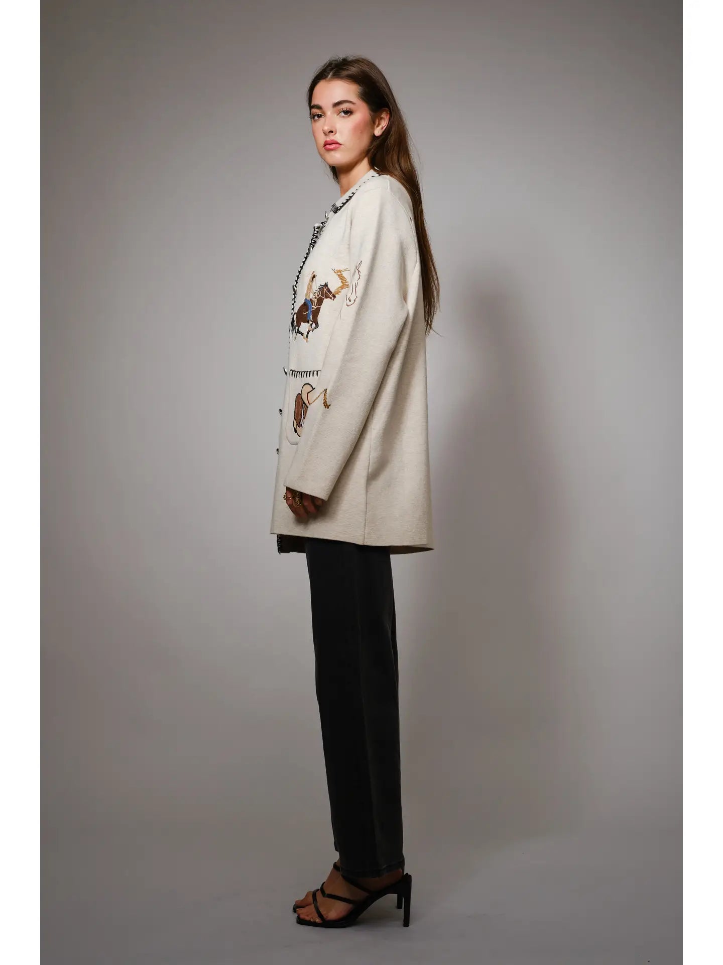 Side view of a woman wearing a cream-colored jacket with western embroidered designs on a gray background
